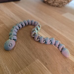 Op de afbeelding: Een flexibel, geleed slangen speelgoed in pastelkleuren blauw, roze en groen. Het speelgoed is gemaakt van verbonden segmenten, waardoor het kan buigen en bewegen. De slang ligt op een houten oppervlak.