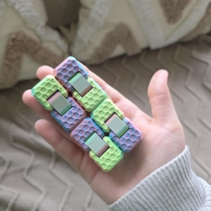 Może przedstawiać: Pastelowa zabawka typu fidget z wzorem plastra miodu trzymana jest w dłoni. Zabawka charakteryzuje się połączeniem odcieni zieleni, różu, błękitu i fioletu. Zabawka składa się z kilku połączonych bloków z metalowym środkiem.