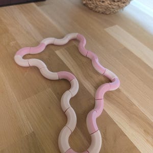 Pode incluir: Um brinquedo segmentado rosa e branco, semelhante a uma cobra, está sobre uma superfície de madeira. O brinquedo é composto por peças curvas e interligadas, alternando entre rosa claro e branco. Uma cesta tecida e uma caixa marrom escura estão no fundo.