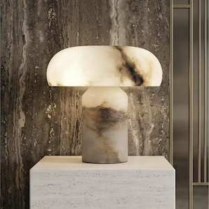 May include: A modern table lamp with a mushroom-shaped shade. The lamp features a cylindrical base and a rounded top, both with a marbled appearance in shades of white, brown, and gray. The lamp is placed on a white stone surface.