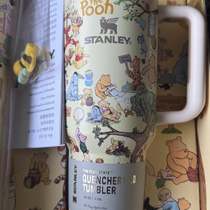 Könnte beinhalten: Ein Stanley Quencher H2.0 Becher mit Winnie Puuh-Figuren. Der 1,18L Becher hat einen cremefarbenen Hintergrund mit Illustrationen von Puuh, Ferkel, Tiger und I-Aah. Er hat einen weißen Griff und einen goldenen Rand.