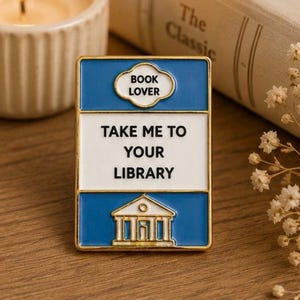 May include: A rectangular enamel pin with a gold-toned metal border. The design features a blue and white colour scheme with the text "BOOK LOVER" in a cloud-shaped frame, followed by the text "TAKE ME TO YOUR LIBRARY" and a white building icon.