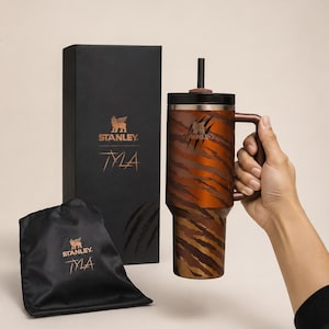 May include: A Stanley Tyla travel mug with a tiger stripe design in shades of brown and orange. The mug has a handle and a black lid with a straw. A black box and a black drawstring bag with the Stanley logo and "Tyla" text are also in the image.