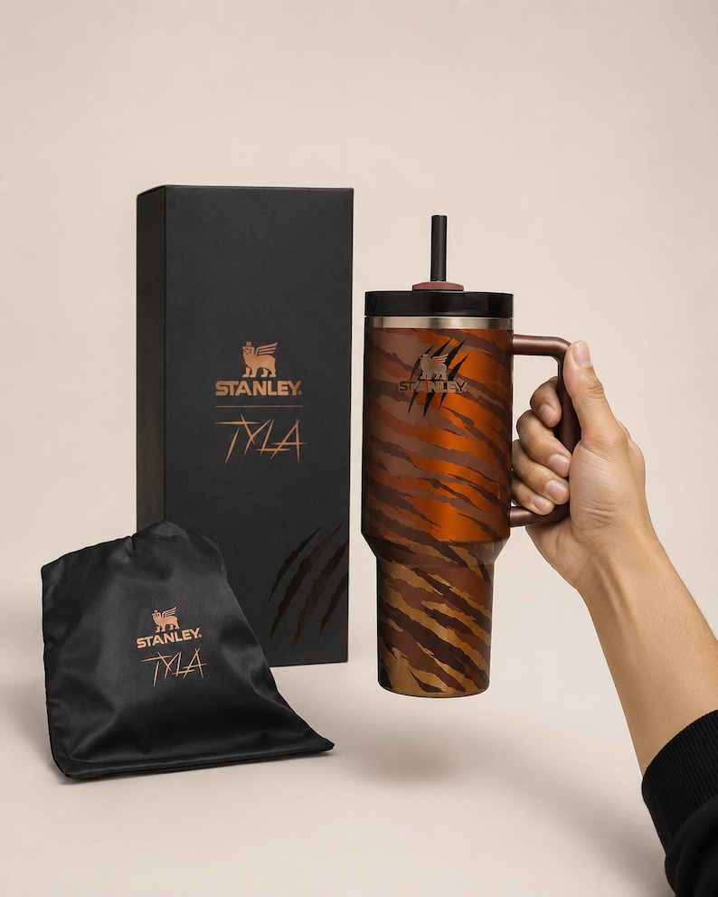 May include: A Stanley Tyla travel mug with a tiger stripe design in shades of brown and orange. The mug has a handle and a black lid with a straw. A black box and a black drawstring bag with the Stanley logo and "Tyla" text are also in the image.