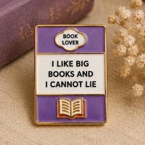 May include: A rectangular enamel pin with gold-tone metal edges. The design features a purple top section with a white cloud-shaped label reading BOOK LOVER, a white centre with the text I LIKE BIG BOOKS AND I CANNOT LIE, and a purple base with an open book.