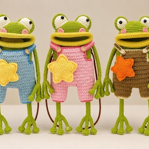 May include: Three handmade crochet frog bags with wide mouths and long limbs. Each frog wears overalls in blue, pink, or brown, featuring a yellow or orange star appliqué on the chest and small buttons on the straps. The bags have brown handles.