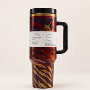 May include: A stainless steel travel mug with a black handle and lid. The mug features a red and gold tiger stripe pattern. A white label on the mug provides information about the product. The mug is designed to keep drinks cold for 11 hours and hot for 7 hours.