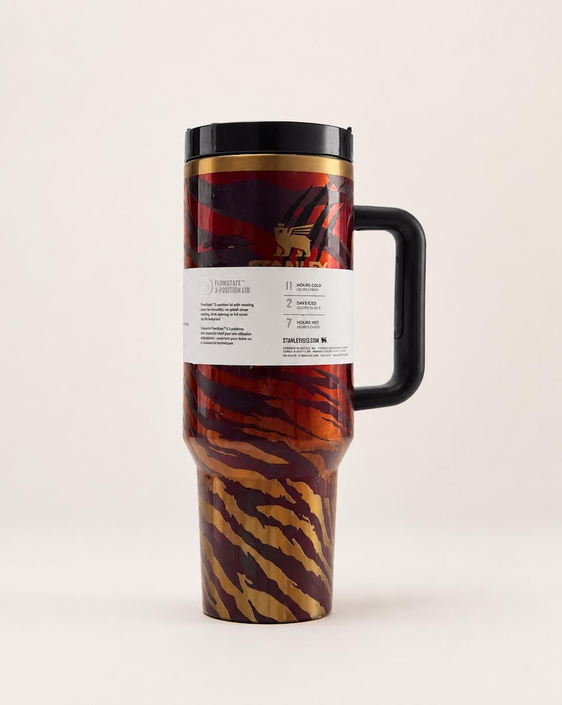 May include: A stainless steel travel mug with a black handle and lid. The mug features a red and gold tiger stripe pattern. A white label on the mug provides information about the product. The mug is designed to keep drinks cold for 11 hours and hot for 7 hours.