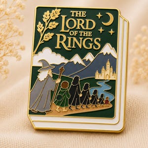 May include: A rectangular enamel pin shaped like a book with a dark green cover. The text THE LORD OF THE RINGS is in gold. It features a gold wizard, a group of figures walking on a path toward a castle, mountains, a crescent moon, and stars.