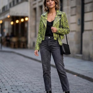 Könnte beinhalten: Eine Frau trägt eine limettengrüne Jacke mit Schlangenmuster, ein schwarzes Oberteil und graue Jeans. Sie trägt schwarze Stiefel und eine schwarze Tasche. Goldene Creolen und eine goldene Halskette ergänzen das Outfit.