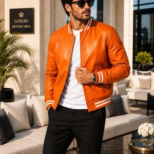 May include: An orange leather varsity jacket featuring a white ribbed collar, cuffs, and hem with double white stripes. The jacket has a snap-button front closure and is worn over a plain white t-shirt paired with black trousers and dark sunglasses.