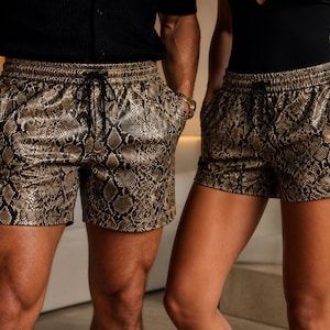 May include: A pair of shorts featuring a metallic gold and black snakeskin pattern. The shorts have an elasticated waistband with a black drawstring cord and two side pockets. The material has a glossy, reflective finish throughout the garment.