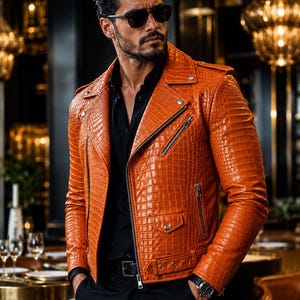 May include: A vibrant orange leather motorcycle jacket featuring a textured crocodile skin pattern, silver-toned metal zips, snap button lapels, and a buckled waist belt, worn over a black button-down shirt with black trousers and dark sunglasses.