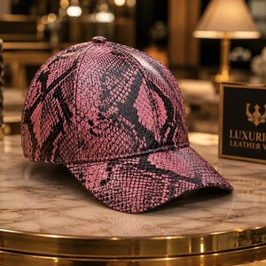 May include: A baseball cap featuring a vibrant pink and black snakeskin pattern. The cap has a structured crown, a curved brim, and a small button at the top center. The design covers the entire exterior surface of the cap in a repeating scale texture.