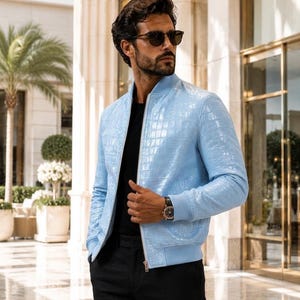 May include: A light blue bomber jacket with a textured crocodile skin pattern, featuring a front zip fastening, ribbed cuffs, and a ribbed hem. The jacket is worn over a plain black shirt and black trousers, paired with dark-tinted sunglasses.