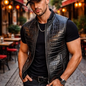 Puede incluir: Un chaleco de cuero negro con un patrón de piel de cocodrilo, usado sobre una camiseta negra. La persona también lleva una gorra negra, un reloj y una cadena plateada. El fondo muestra una calle adoquinada y edificios.