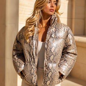 May include: A tan and brown snakeskin-patterned puffer jacket with a high collar and a front zipper. The jacket is worn over a light-colored top and pants. The model is wearing a gold necklace.
