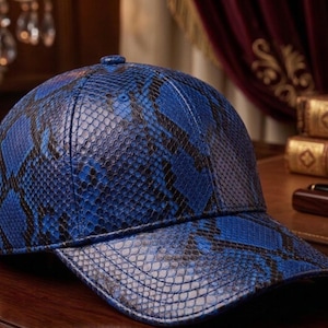 Puede incluir: Gorra de béisbol azul y negra con estampado de piel de serpiente. La gorra tiene una visera curva y una correa ajustable con una hebilla dorada. El sombrero está hecho de un material texturizado y se muestra sobre una superficie de madera.