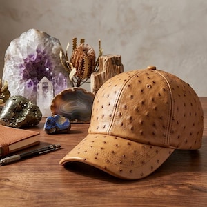 May include: A tan baseball cap with a textured surface, featuring a curved brim and a button on top. The cap is made of a leather-like material. A notebook, pencil, and various crystals are in the background.