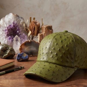 May include: An olive green baseball cap with a textured surface, resting on a wooden table. The cap is the focal point, with a notebook, pen, and various crystals and stones in the background. The cap's design is a key feature.