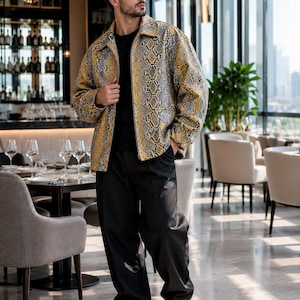 Puede incluir: Una chaqueta de manga larga con un patrón de piel de serpiente en amarillo y gris, usada sobre una camiseta negra lisa y combinada con pantalones negros holgados y zapatos de cuero negro. La chaqueta tiene cuello y cremallera frontal.