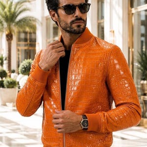 Può includere: Un giubbotto bomber arancione brillante caratterizzato da una texture lucida in rilievo effetto pelle di coccodrillo. La giacca presenta colletto, polsini e orlo a coste, una chiusura frontale con zip e una silhouette aderente ed elegante.