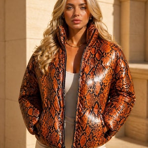 May include: A shiny, orange and black snakeskin-patterned puffer jacket with a high collar and a full-length zipper. The jacket is worn over a light-colored top.