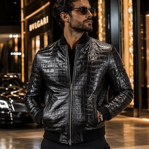 Può includere: Un giubbotto bomber nero caratterizzato da una texture con stampa a coccodrillo, una chiusura frontale con zip integrale e polsini e orlo a coste neri. Il capo è indossato sopra una maglia nera e presenta maniche lunghe e tasche laterali.