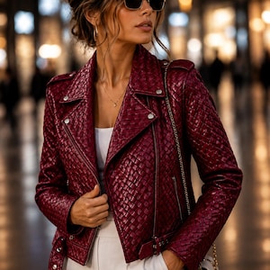 May include: A deep burgundy biker jacket with a woven leather texture, featuring silver-toned metal hardware, a notched lapel, and an asymmetrical front zip closure. The jacket is worn over a plain white top and paired with light-coloured trousers.