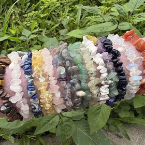 Puede incluir: Una colección de pulseras de virutas de piedras preciosas en varios colores, incluyendo marrón, blanco, azul, amarillo, rosa, verde, morado, negro y naranja. Las pulseras se exhiben sobre una superficie rosa contra un fondo de follaje verde.