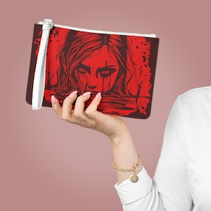 Puede incluir: Un clutch rectangular con un diseño gráfico rojo y marrón. El diseño presenta el rostro de una mujer con lágrimas y una correa de muñeca blanca. El clutch tiene una cremallera blanca y un borde marrón.