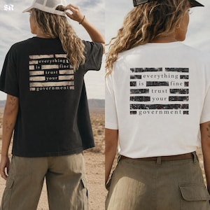 Può includere: Due magliette, una nera e una bianca, con la scritta "everything is fine trust your government" stampata sul retro in un design rettangolare. Le magliette sono indossate da una persona in un ambiente desertico.