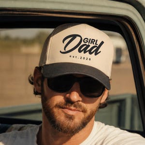 May include: A beige and black baseball cap with the words "Girl Dad" and the est. 2026 printed on the front. The cap has a curved brim and is worn by a person wearing sunglasses.