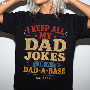 Könnte beinhalten: Schwarzes T-Shirt mit dem Text "I KEEP ALL MY DAD JOKES IN A DAD-A-BASE est. 2026" in Rot, Beige und Blau. Das Design hat einen Vintage-Look mit Sternen und dekorativen Linien. Rundhalsausschnitt und kurze Ärmel.