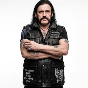 Puede incluir: Retrato de un hombre con pelo largo y negro y un gran bigote, vistiendo un chaleco de cuero negro con parches. El chaleco tiene el texto "Everything Louder Than Everything Else" y un gráfico de calavera. También lleva una camiseta negra y un collar con un colgante de cruz.