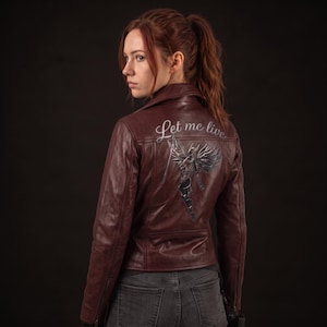 May include: A dark burgundy leather jacket featuring a pointed collar, long sleeves, and zippered cuffs. The back displays a graphic of a winged figure in armor with the script text Let me live printed in white above the illustration.