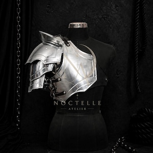 May include: A silver metal pauldron and breastplate armor piece featuring layered shoulder plates and a curved chest section secured with dark leather straps. The armor is displayed on a black mannequin with the text NOCTELLE ATELIER visible.