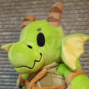 May include: A plush green dragon toy with brown horns, yellow wings, and a smiling face. The dragon wears a tan outfit with brown accents. The toy is soft and cuddly, perfect for children.