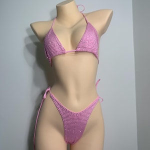 Puede incluir: Conjunto de bikini rosa, con top triangular y braguita de corte alto, totalmente adornado con pequeños y brillantes pedrería. El bikini se exhibe en un maniquí, mostrando el top halter y la braguita con lazos laterales. El color es un rosa claro vibrante.