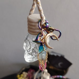 May include: A clear skull-shaped car air freshener with a rainbow-colored crystal pendant wrapped in gold wire. A gold crescent moon charm and a small witch hat charm hang from the pendant. The air freshener is suspended by a beige and gold rope.