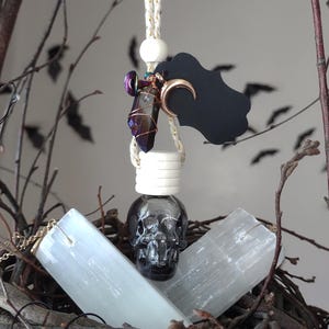 May include: A black skull-shaped bottle with a crystal pendant hanging from it. The bottle is surrounded by white selenite crystals and a black tag with a crescent moon.