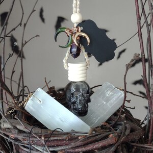 May include: A black skull-shaped glass bottle with a white stopper hangs from a gold chain with green and gold crescent moon charms. The bottle is nestled in a brown twig nest with two white selenite sticks.