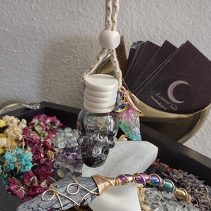 May include: A car air freshener with a skull-shaped glass bottle, a wooden cap, and a decorative charm. The freshener hangs from a beige and gold rope. A black kyanite wand with gold wire wrapping and iridescent beads is also visible.