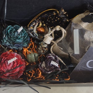 May include: A black box filled with Halloween themed items, including three floral candles, a small bottle with a skull inside, a small green pumpkin, a black velvet bag with stars and a crescent moon, and a burlap bag with a white stick inside. The box is lined with black and orange shredded paper. A business card with a crescent moon logo and the text "Enchanted Handmade" is visible in the bottom right corner.