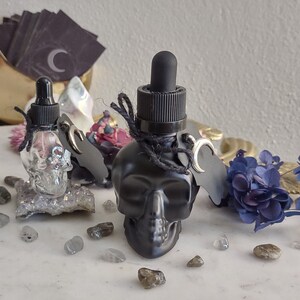 May include: Two black glass bottles with skull-shaped containers and silver droppers. The bottles are tied with black string and have a silver crescent moon charm. The bottles are sitting on a white surface with small grey stones.
