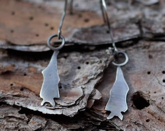 Puede incluir: Un par de pendientes de plata con un diseño abstracto único. Los pendientes cuelgan de una superficie rugosa y texturizada.