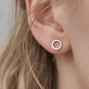 May include: A close-up of a person's ear with a small, silver circle earring. The earring is a simple, minimalist design, and the focus is on the jewelry. The earring is a stud style, and the metal appears to be polished.