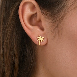 Peut inclure: Une boucle d'oreille à clou dorée en forme de palmier. Le bijou présente un tronc central avec plusieurs palmes incurvées s'étendant depuis le sommet, créant la silhouette d'un palmier. La pièce est polie avec une finition lisse et brillante.