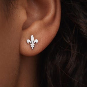 May include: A close-up of a silver fleur-de-lis stud earring on an ear. The earring is a small, detailed representation of the heraldic symbol. The earring is set against the skin of the ear and surrounding area.