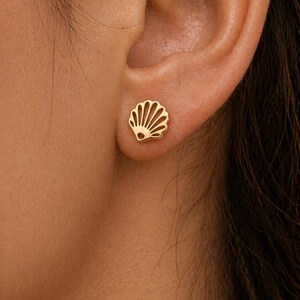 Peut inclure: Une boucle d'oreille à clou dorée en forme de coquillage. Le bijou présente un design ajouré avec des lignes rayonnantes imitant les crêtes naturelles d'une coquille Saint-Jacques. La pièce est petite et repose à plat sur le lobe de l'oreille.